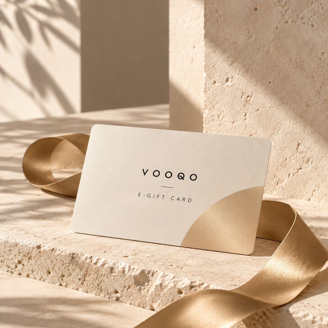 A VOOQO e-gift card with a minimalist cream and gold design, styled on a textured stone pedestal with a flowing gold ribbon under warm, natural shadows.