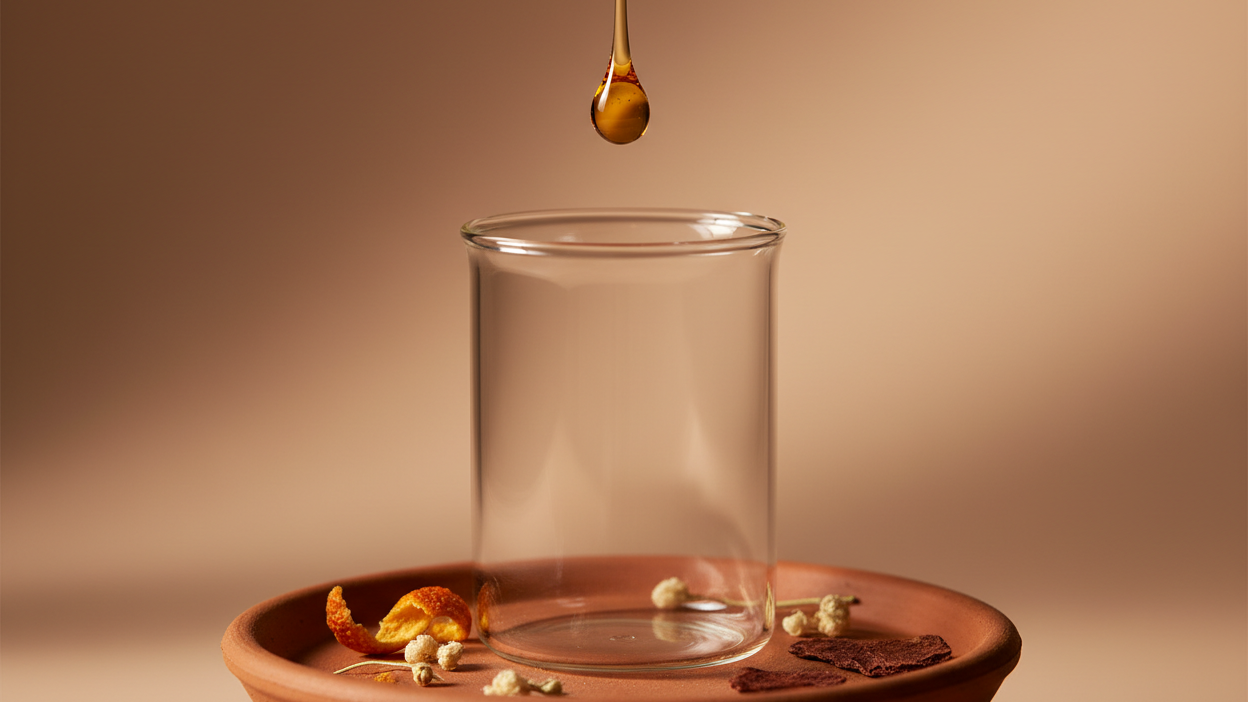 Glass cup on a terracotta dish with a droplet of liquid above it on a brown background
