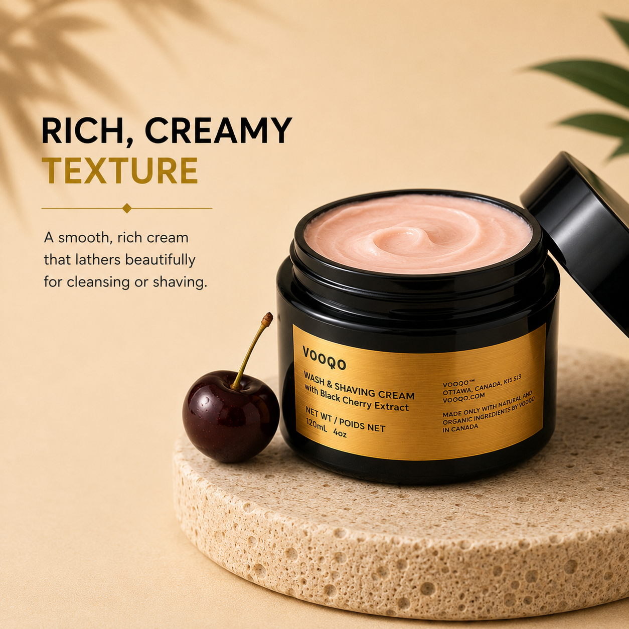 A jar of VOOQO wash and shaving cream with black cherry extract, shown open with the cream visible, on a textured stone slab with a cherry prop. The background is a warm beige with text describing the rich, creamy texture.